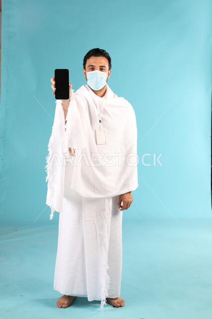 الإلتزام بالاحترازات والاجراءات الوقائية بإرتداء الكمامة, عرض شاشة سوداء فارغة، بورتريه لرجل عربي خليجي سعودي يرتدي لباس الاحرام يقف باستقامة يمسك الهاتف المحمول، اداء مناسك الحج والعمرة، تطبيقات مواعيد الأذان والصلاة، صورة الجسم بالطول الكامل، خلفية زرقاء