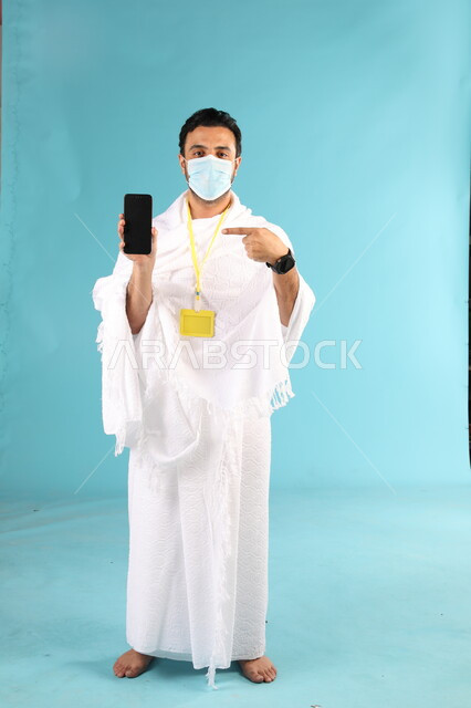 الإلتزام بالإجراءات الوقائية بإرتداء الكمامة, عرض شاشة سوداء فارغة، بورتريه لرجل عربي خليجي سعودي يرتدي لباس الاحرام يقف باستقامة يمسك الهاتف المحمول يشير اليه باصبع السبابة، اداء مناسك الحج والعمرة، تطبيقات مواعيد الأذان والصلاة، صورة لكامل الجسم، خلفية زرقاء