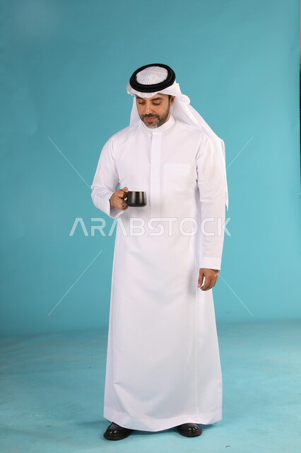 احتساء المشروبات الساخنه, الاستمتاع بمذاق القهوة العربية، بورتريه لرجل عربي خليجي سعودي يرتدي الغترة والثوب التقليدي يحمل بيده كوب اسود, صورة الجسم بالطول الكامل، خلفية زرقاء