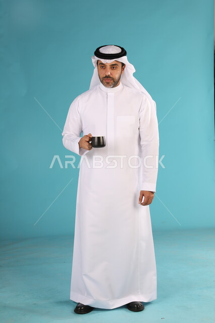 احتساء المشروبات الساخنه, الاستمتاع بمذاق القهوة العربية، بورتريه لرجل عربي خليجي سعودي يرتدي الغترة والثوب التقليدي يحمل بيده كوب اسود, صورة الجسم بالطول الكامل، خلفية زرقاء