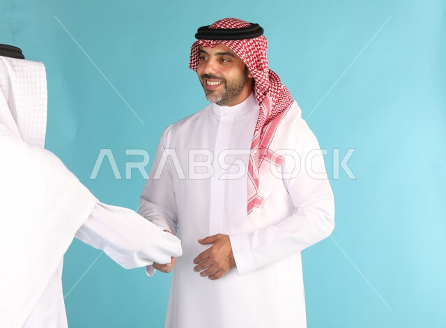 عقد الاتفاقات والصفقات التجارية بين رجال الأعمال، بورتريه مقرب لرجل عربي خليجي سعودي مبتسم يرتدي الشماغ والثوب التقليدي يقوم بمصافحة شخص اخر، الترحيب والاستقبال بتعابير البشاشة والسرور، خلفية زرقاء