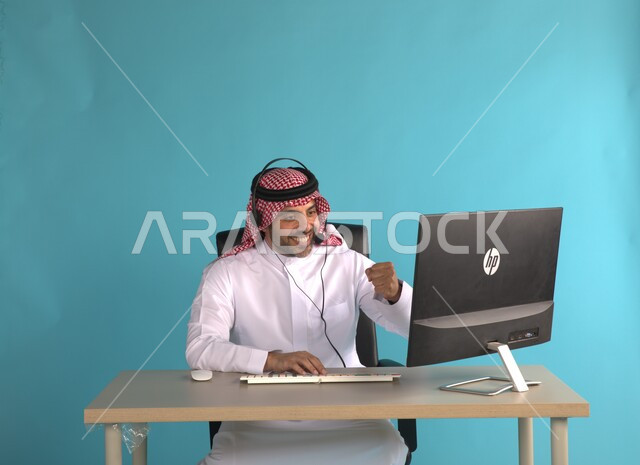 مفهوم خدمة العملاء، الرد والاجابة على الاستفسارات، اجراء اجتماع عمل عن بعد باستخدام الحاسوب، بورتريه لرجل اعمال عربي خليجي سعودي مبتسم يرتدي الشماغ والثوب التقليدي يجلس في مكتبه يضع سماعة الراس، مهن ووظائف مكتبية سعودية، خلفية زرقاء