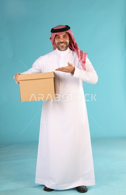 خدمات التوصيل السريع، بورتريه لرجل عربي سعودي خليجي مبتسم يرتدي الشماغ والثوب التقليدي يحمل صندوق كرتون كبير ويشير اليه، تسليم الطلبات والبضائع للعملاء, صورة الجسم بالطول الكامل، خلفية زرقاء