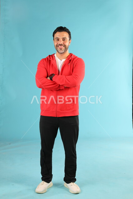 الوقوف باستقامة وايدي متقاطعة بايماءات وتعابير السرور والسعادة، الرياضات السعودية الشبابية، ممارسة الهوايات الرياضية، بورتريه لمدرب عربي خليجي سعودي مبتسم يرتدي الملابس الرياضية ينظر الى الكاميرا، صورة الجسم بالطول الكامل، خلفية زرقاء