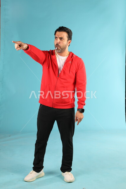 استخدام الصفارة للتنبيه والتحذير، النظر والاشارة الى شيء ما، الرياضات السعودية الشبابية، ممارسة الهوايات الرياضية، بورتريه لمدرب عربي خليجي سعودي يرتدي الملابس الرياضية يقوم بالتصفير يشير باصبع السبابة، صورة الجسم بالطول الكامل، خلفية زرقاء