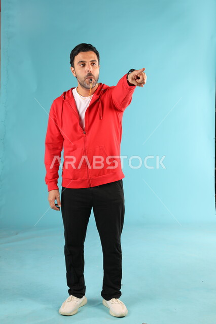 النظر والاشارة الى شيء ما، الرياضات السعودية الشبابية، ممارسة الهوايات الرياضية، استخدام الصفارة للتنبيه والتحذير، بورتريه لمدرب عربي خليجي سعودي يرتدي الملابس الرياضية يقوم بالتصفير يشير باصبع السبابة، صورة الجسم بالطول الكامل، خلفية زرقاء