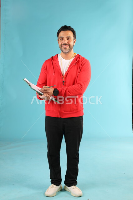 ممارسة الهوايات الرياضية، تعابير وايماءات السرور والسعادة، الوقوف باستقامة والنظر الى الكاميرا، الرياضات السعودية الشبابية، بورتريه لمدرب عربي خليجي سعودي مبتسم يرتدي الملابس الرياضية يقوم بكتابة الملاحظات حول الفريق، صورة الجسم بالطول الكامل، خلفية زرقاء