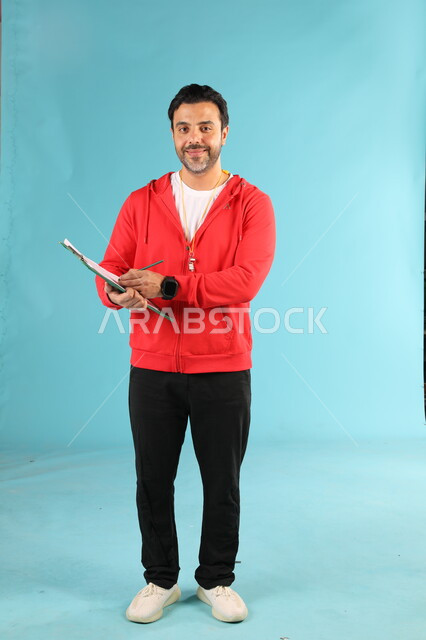 ممارسة الهوايات الرياضية، تعابير وايماءات السرور والسعادة، الوقوف باستقامة والنظر الى الكاميرا، الرياضات السعودية الشبابية، بورتريه لمدرب عربي خليجي سعودي مبتسم يرتدي الملابس الرياضية يقوم بكتابة الملاحظات حول الفريق، صورة الجسم بالطول الكامل، خلفية زرقاء