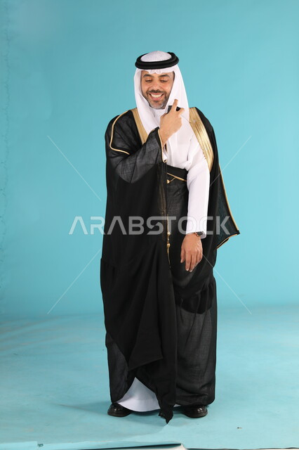 التطيب بالعطور والروائح الفاخرة، استخدام الزيوت العطرية الطبيعية، بورتريه لرجل عربي خليجي سعودي يرتدي البشت والغترة التقليدية يقوم برش العطر، مراسم الأعراس والزواج في السعودية، مفهوم الأناقة والاهتمام بالمظهر الخارجي، صورة الجسم بالكامل, خلفية زرقاء