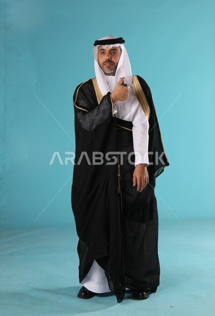 التطيب بالعطور والروائح الفاخرة، استخدام الزيوت العطرية الطبيعية، بورتريه لرجل عربي خليجي سعودي يرتدي البشت والغترة التقليدية يقوم برش العطر، مراسم الأعراس والزواج في السعودية، مفهوم الأناقة والاهتمام بالمظهر الخارجي، صورة الجسم بالكامل, خلفية زرقاء