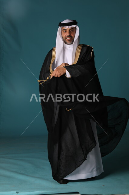 مفهوم الاستقرار وتكوين الأسرة, بورتريه لعريس عربي خليجي سعودي يرتدي البشت الأسود والثوب التقليدي يتظر إلى شيء ما, الاهتمام بالمظهر الخارجي, مراسم الأعياد والزواج في السعودية, صورة الجسم بالطول الكامل, خلفية زرقاء