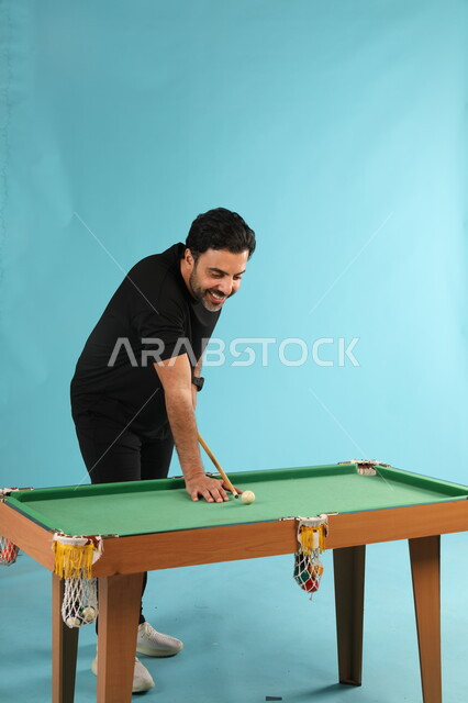 قضاء الوقت الممتع بممارسة الهوايات الترفيهيه, التسلية والترفيه في أوقات الفراغ, بورتريه لشاب عربي خليجي سعودي مبتسم يرتدي الزي الكاجوال يلعب البلياردو (السنوكر), التركيز والهدوء والتصويب, أنشطة شبابية ممتعة, صورة الجسم بالطول الكامل, خلفية زرقاء