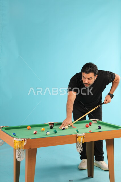 التركيز والهدوء والتصويب, قضاء الوقت الممتع بممارسة الهوايات الترفيهيه, التسلية والترفيه في أوقات الفراغ, بورتريه لشاب عربي خليجي سعودي يرتدي الزي الكاجوال يلعب البلياردو (السنوكر), أنشطة شبابية ممتعة, صورة الجسم بالطول الكامل, خلفية زرقاء