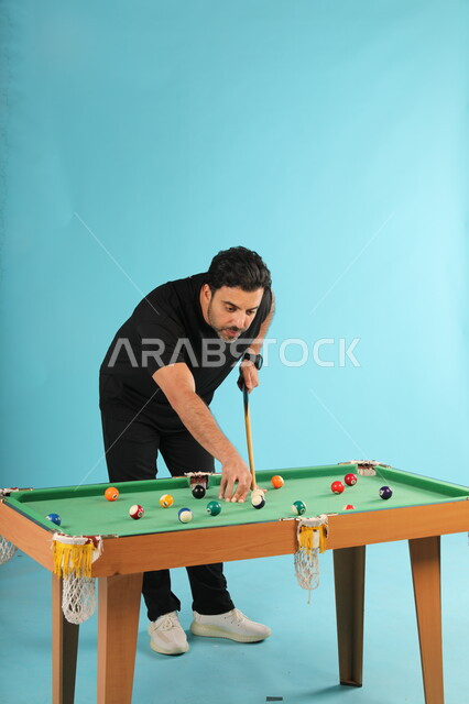 قضاء الوقت الممتع بممارسة الهوايات الترفيهيه, التسلية والترفيه في أوقات الفراغ, بورتريه لشاب عربي خليجي سعودي يرتدي الزي الكاجوال يلعب البلياردو (السنوكر), التركيز والهدوء والتصويب, أنشطة شبابية ممتعة, صورة الجسم بالطول الكامل, خلفية زرقاء