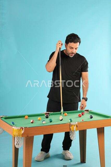 التسلية والترفيه وقضاء الوقت الممتع, أنشطة وهوايات شبابية, الاستمتاع والشغف بممارسة لعبة السنوكر, بورتريه لشاب عربي خليجي سعودي يرتدي الزي الكاجوال يلعب البلياردو, صورة الجسم بالطول الكامل, خلفية زرقاء