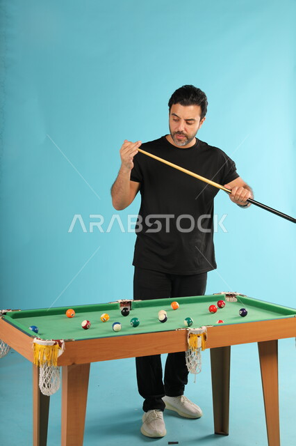 Youth activities and hobbies, enjoyment and passion for playing snooker, portrait of a Saudi Gulf Arab young man wearing casual clothes playing billiards, entertainment and spending a good time, full-length body image, blue background