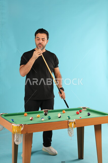 التسلية والترفيه وقضاء الوقت الممتع, أنشطة وهوايات شبابية, الاستمتاع والشغف بممارسة لعبة السنوكر, بورتريه لشاب عربي خليجي سعودي يرتدي الزي الكاجوال يلعب البلياردو, صورة الجسم بالطول الكامل, خلفية زرقاء