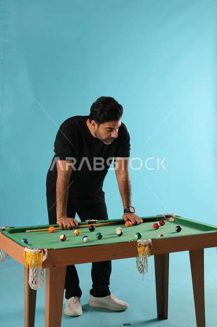 Concentration, calmness and aiming, spending enjoyable time practicing recreational hobbies, amusement and leisure time, portrait of a Saudi Gulf Arab young man wearing a casual outfit playing billiards (snooker), fun youth activities, full-length body portrait, blue background