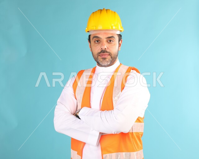 مفهوم الهندسة والانشاءات المعمارية، بورتريه مقرب لمهندس عربي خليجي سعودي يرتدي الخوذه و سترة الحماية ينظر إلى الكاميرا، الوقوف بإيماءات أيدي متقاطعه، العمل في القطاع الهندسي، مهن ووظائف سعودية، الإعمار بسواعد أبناء الوطن، خلفية زرقاء
