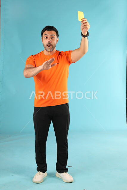 رفع البطاقة الصفراء للإنذار, بورتريه لمدرب عربي خليجي سعودي يرتدي الزي الرياضي يقوم بالتصفير, استخدام الصفارة للتنبيه والتحذير, ممارسة الرياضات الشبابية الممتعه, صورة الجسم بالطول الكامل, خلفية زرقاء