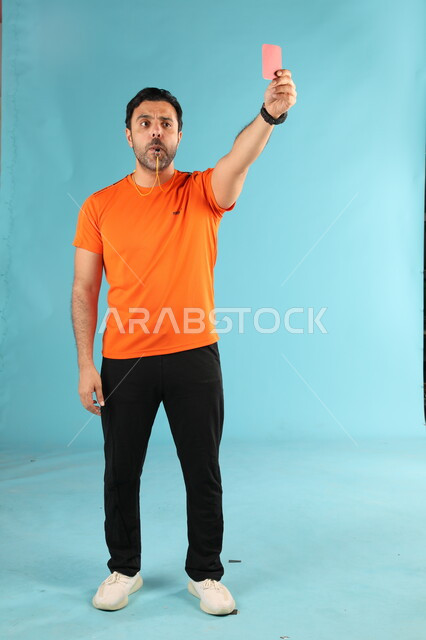 Expulsion from the match using a red card, portrait of a Saudi Gulf Arab coach wearing a sports uniform, blowing the siren and warning, passion for youth sports activities, fun, entertainment and spending a good time, full-length body image, blue background