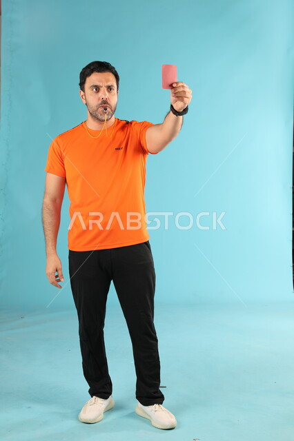 الطرد من المباراة باستخدام البطاقة الحمراء, بورتريه لمدرب عربي خليجي سعودي يرتدي الزي الرياضي يقوم بالتصفير عبر صفارة الانذار والتحذير, الشغف بالأنشطة الرياضية الشبابية, المتعه والتسلية وقضاء الوقت الممتع, صورة الجسم بالطول الكامل, خلفية زرقاء