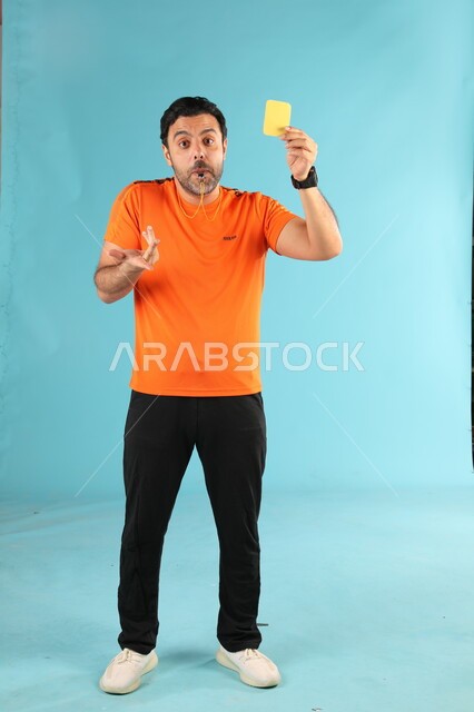 رفع البطاقة الصفراء للإنذار, بورتريه لمدرب عربي خليجي سعودي يرتدي الزي الرياضي يقوم بالتصفير, استخدام الصفارة للتنبيه والتحذير, ممارسة الرياضات الشبابية الممتعه, صورة الجسم بالطول الكامل, خلفية زرقاء