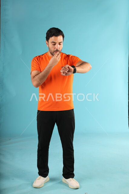 تحديد الوقت للجولات في المباريات, بورتريه لمدرب عربي خليجي سعودي يرتدي الزي الرياضي ينظر إلى الساعة, ممارسة الهوايات والأنشطة الشبابية, استخدام الصفارة للتحذير والتنبيه, صورة الجسم بالطول الكامل, خلفية زرقاء