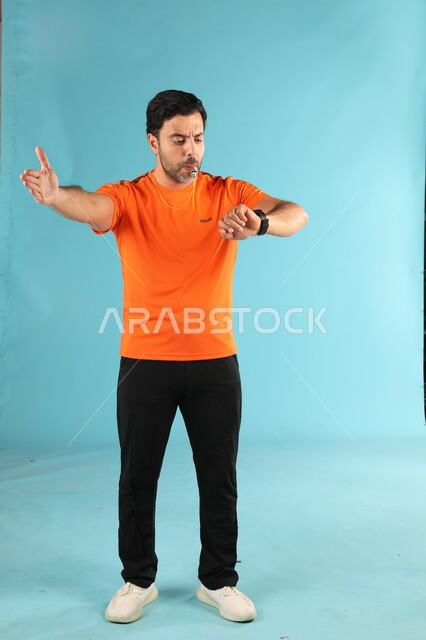 Setting the time for rounds in matches, a portrait of a Saudi Gulf Arab coach wearing a sports uniform looking at the clock, practicing hobbies and youth activities, using the whistle to warn and alert, full-length body portrait, blue background