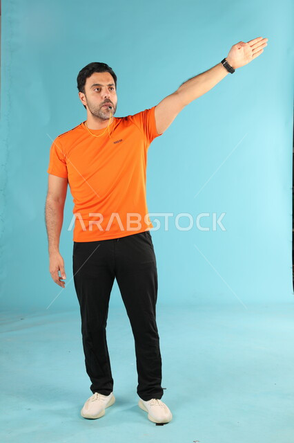 النظر والاشارة الى شيء ما، الرياضات السعودية الشبابية، ممارسة الهوايات الرياضية، استخدام الصفارة للتنبيه والتحذير، بورتريه لمدرب عربي خليجي سعودي يرتدي الملابس الرياضية يقوم بالتصفير يرفع يده للأعلى، صورة الجسم بالطول الكامل، خلفية زرقاء
