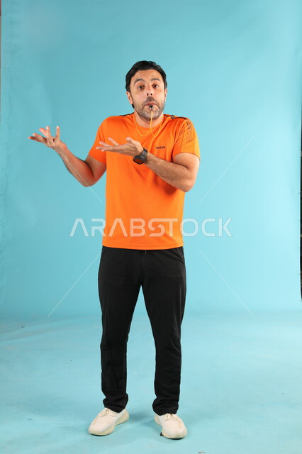 الوقوف بإيماءات الاستفهام والسؤال، الرياضات السعودية الشبابية، الشغف بممارسة الهوايات الرياضية، استخدام الصفارة للتنبيه والتحذير، بورتريه لمدرب عربي خليجي سعودي يرتدي الملابس الرياضية يقوم بالتصفير، صورة الجسم بالطول الكامل، خلفية زرقاء