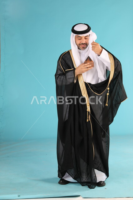 مفهوم الأناقة والاهتمام بالمظهر الخارجي, التطيب بالعطور والروائح الفاخرة، استخدام الزيوت العطرية الطبيعية، بورتريه لرجل عربي خليجي سعودي يرتدي البشت والغترة التقليدية يقوم برش العطر، مراسم الأعراس والزواج في السعودية، صورة الجسم بالكامل, خلفية زرقاء