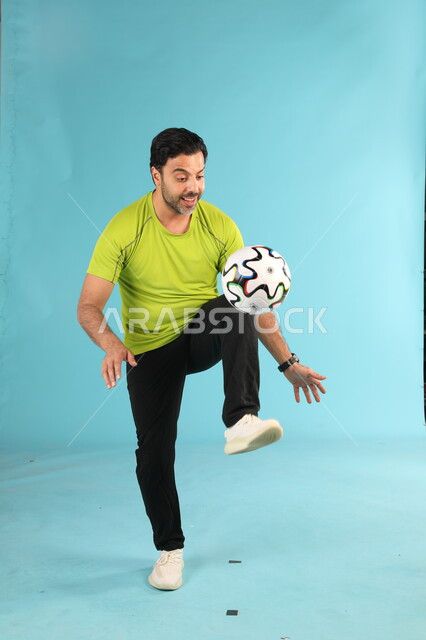 هواية اللعب بالكرة, التدريب الرياضي والاستعداد لبدء المباراة, بورتريه للاعب عربي خليجي سعودي يرتدي الزي الرياضي يقوم بالحركات الرياضية الشغف بممارسة الهوايات الشبابية, التسلية والترفيه بالألعاب الجماعية, صورة الجسم بالطول الكامل, خلفية زرقاء