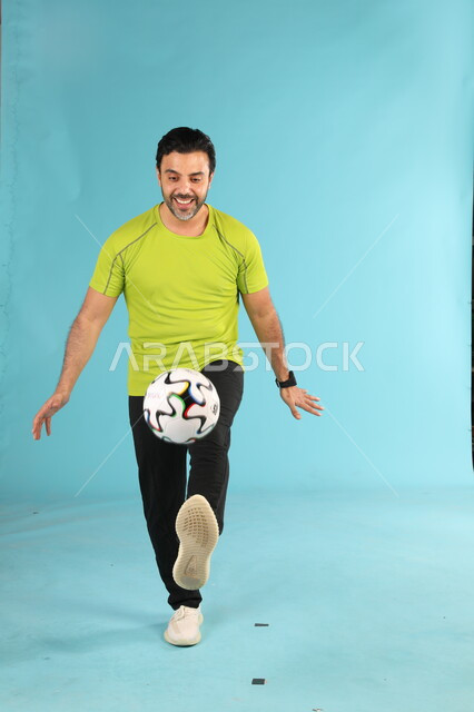التدريب الرياضي والاستعداد لبدء المباراة, هواية اللعب بالكرة, بورتريه للاعب عربي خليجي سعودي يرتدي الزي الرياضي يقوم بالحركات الرياضية الشغف بممارسة الهوايات الشبابية, التسلية والترفيه بالألعاب الجماعية, صورة الجسم بالطول الكامل, خلفية زرقاء