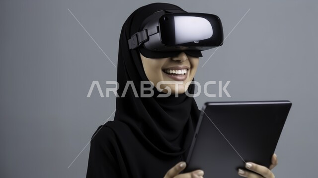 Expressions of happiness with advanced technologies, the use of technology and modern technical devices, a close-up portrait of a veiled Saudi Arabian Gulf woman, smiling, wearing a black abaya, wearing virtual reality glasses and using a portable tablet with gestures of happiness and pleasure, gray background