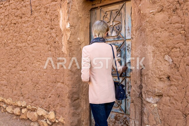  البيوت والمنشآت المبنية على الطراز الطيني القديم في قرية أشيقر الأثرية في محافظة شقراء, معالم وأماكن الجذب السياحي من شتى أنحاء العالم, القرى الشعبية في المملكة العربية السعودية, صورة من الخلف لسائحة غربية تتجول في إحدى الأحياء التراثية الضيقة في النهار