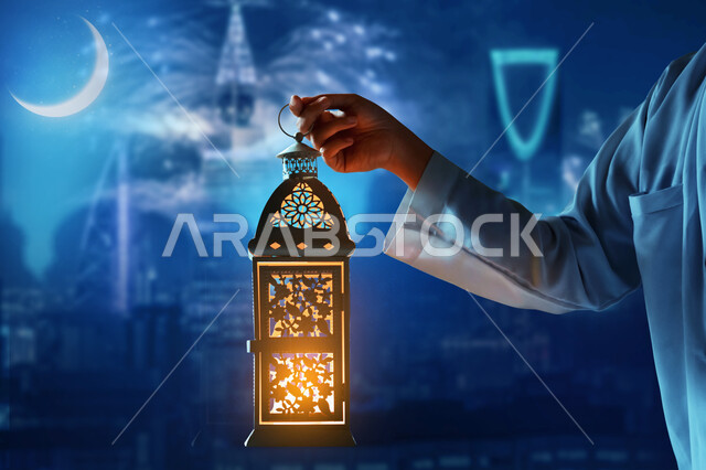 زينة الأعياد والمناسبات الدينية, صورة مقربة ليد رجل عربي خليجي سعودي يرتدي الثوب الأبيض يمسك بيده فانوس معدني مزخرف متوهج, الاستعداد لاستقبال شهر الخير, احتفال المسلمين بقدوم رمضان المبارك, خلفية الأبراج وناطحات السحاب