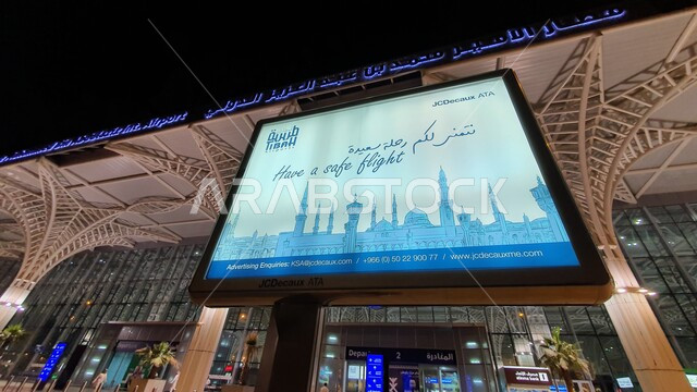 شاشة الكترونية مضاءة مع عبارات وتمنيات السلامة للمسافرين، محطات استقبال ومغادرة السائحين في المدينة المنورة، الفن الهندسي المعماري المتطور لمنشآت السفر الجوية والبرية في المملكة العربية السعودية، الواجهه الخارجية لمطار الأمير محمد بن عبد العزيز الدولي في الليل