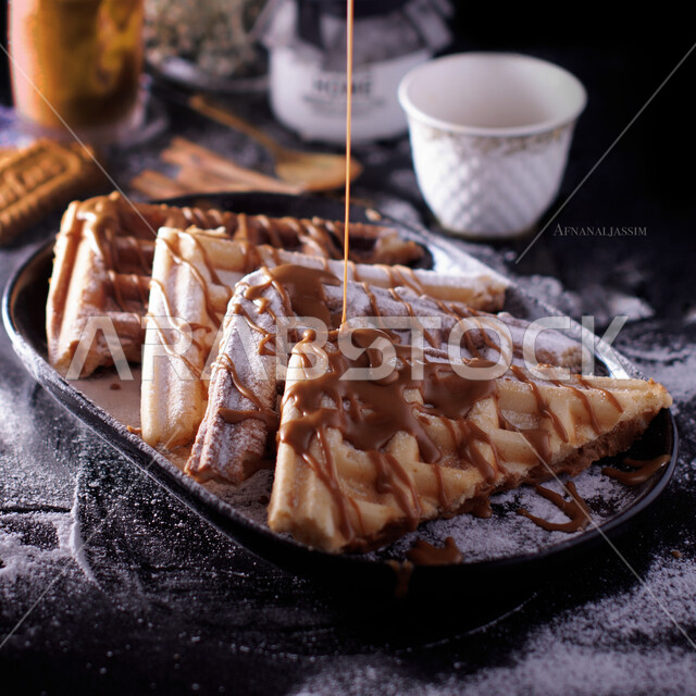 A plate of waffle dessert decorated with liquid chocolate next to a cup of Saudi Arabian coffee, delicious candy pieces, delicious cold desserts, and high-calorie dishes.