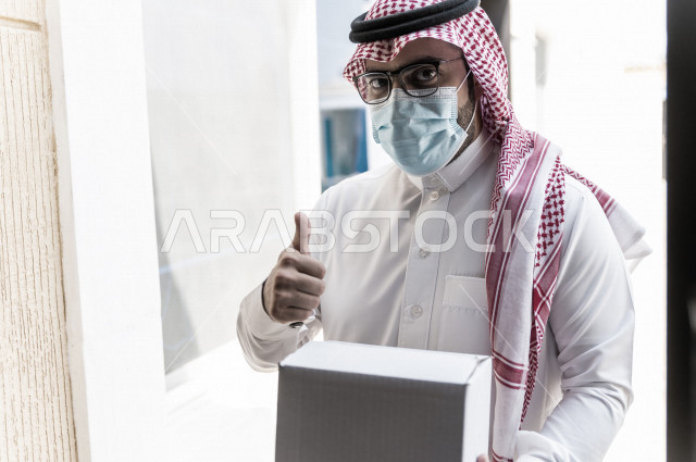 شاب سعودي خليجي يعمل على توصيل الطلبات يرتدي الكمامة ، توصيل صناديق  و حقائب تحتوي على طلبات و مستلزمات لزبونة سعودية لباب البيت ، عمليه شحن و توصيل موثوقة، امتلاك بيت جديد ، النقل و التوصيل 