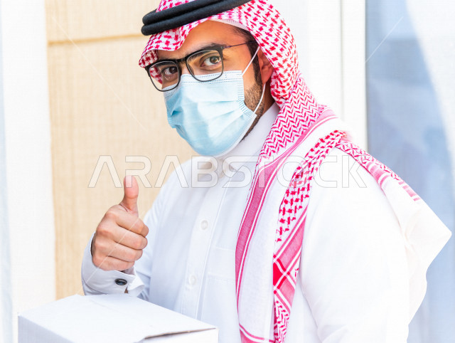 شاب سعودي خليجي يعمل على توصيل الطلبات يرتدي الكمامة ، توصيل صناديق  و حقائب تحتوي على طلبات و مستلزمات لزبونة سعودية لباب البيت ، عمليه شحن و توصيل موثوقة، امتلاك بيت جديد ، النقل و التوصيل 