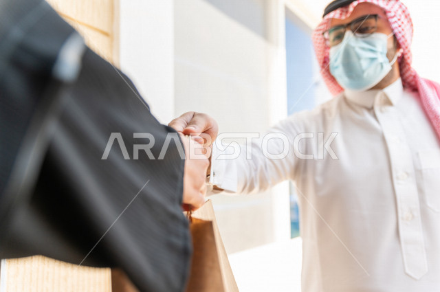 شاب سعودي خليجي يعمل على توصيل الطلبات يرتدي الكمامة ، توصيل صناديق  و حقائب تحتوي على طلبات و مستلزمات لزبونة سعودية لباب البيت ، عمليه شحن و توصيل موثوقة، امتلاك بيت جديد ، النقل و التوصيل 