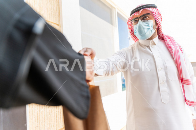 شاب سعودي خليجي يعمل على توصيل الطلبات يرتدي الكمامة ، توصيل صناديق  و حقائب تحتوي على طلبات و مستلزمات لزبونة سعودية لباب البيت ، عمليه شحن و توصيل موثوقة، امتلاك بيت جديد ، النقل و التوصيل 