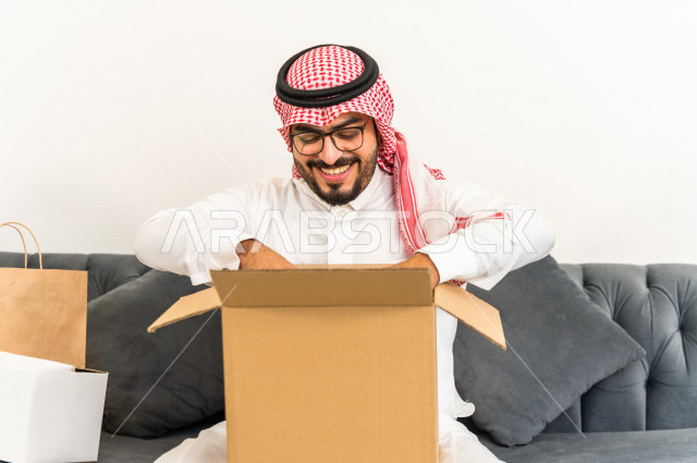 رجل عربي سعودية يجلس على كنبة داخل منزله الجديد ، يحمل بيده كرتونة  بها أغراضه الشخصية ،امتلاك بيت جديد ،ايماءات وجه و يدين تدل على السعادة والانسجام ، الترتيب و التنظيم ، النقل و التوصيل 