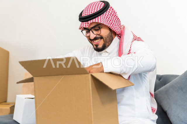 A Saudi-Arab man sits on a sofa inside his new home, carrying a cardboard with his personal belongings in his hand, owning a new house, face and hand gestures that indicate happiness and harmony, arrangement and organization, transportation and delivery
