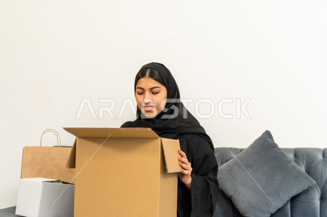 مرأه عربية سعودية تجلس على كنبة داخل منزلها الجديد ، تحمل بيدها كرتونة  بها أغراضها الشخصية ،امتلاك بيت جديد ،ايماءات وجه و يدين تدل على السعادة والانسجام ، الترتيب و التنظيم ، النقل و التوصيل 