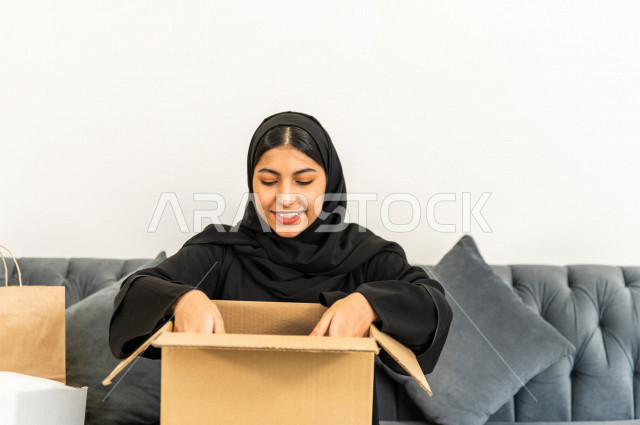مرأه عربية سعودية تجلس على كنبة داخل منزلها الجديد ، تحمل بيدها كرتونة  بها أغراضها الشخصية ،امتلاك بيت جديد ،ايماءات وجه و يدين تدل على السعادة والانسجام ، الترتيب و التنظيم ، النقل و التوصيل 