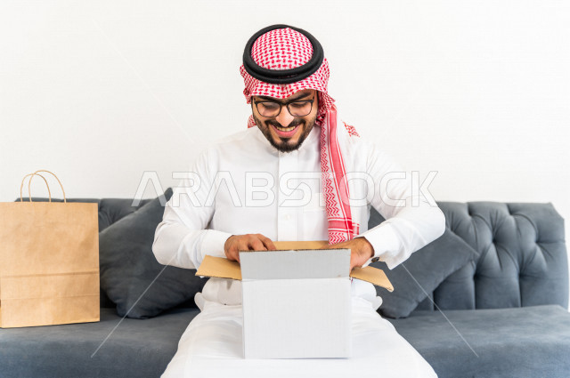A Saudi-Arab man sits on a sofa inside his new home, carrying a cardboard with his personal belongings in his hand, owning a new house, face and hand gestures that indicate happiness and harmony, arrangement and organization, transportation and delivery