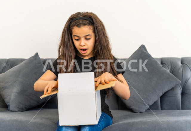 طفلة عربية سعودية تجلس على كنبة داخل منزلها الجديد ، تحمل بيدها كرتونة  بها أغراضها الشخصية ،امتلاك بيت جديد ،ايماءات وجه و يدين تدل على السعادة والانسجام ، الترتيب و التنظيم ، النقل و التوصيل 