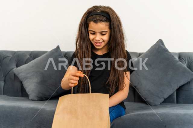 A Saudi Arab girl sits on a sofa inside her new home, carrying a bag containing her personal belongings in her hand, owning a new home, face and hands gestures indicating happiness and harmony, arrangement and organization, transportation and delivery